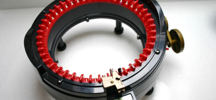 A circular knitting machine with red needles and a black base, placed on a white surface.