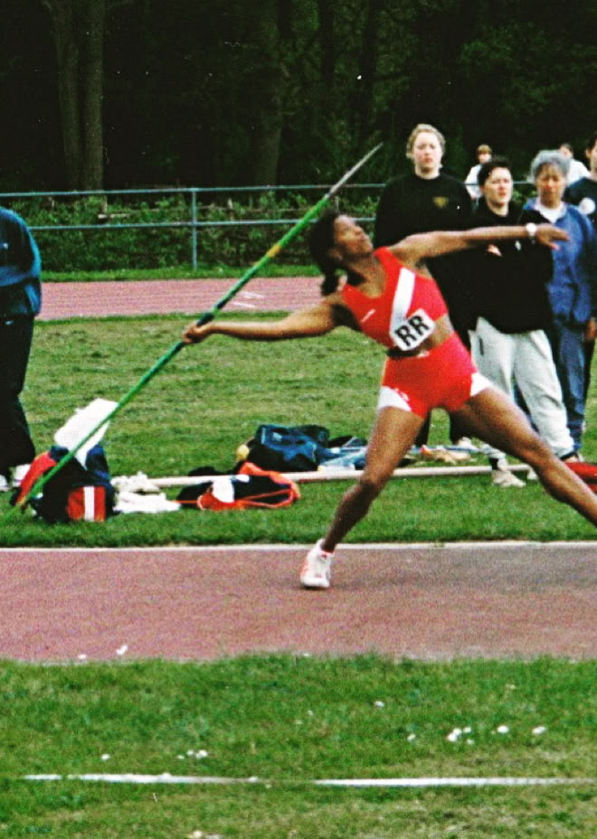 Athlete in red uniform preparing to throw a javelin on a track, with spectators in the background.