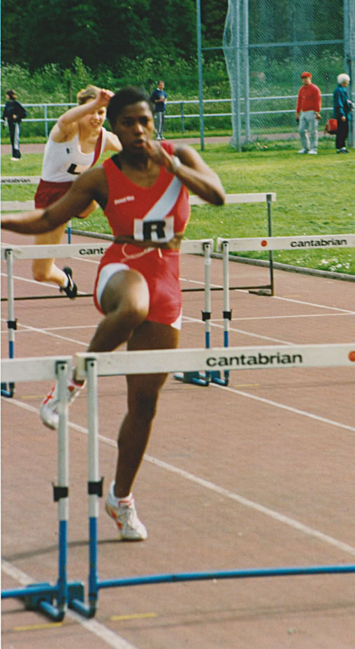 Athlete in red uniform jumping over a hurdle on a track.